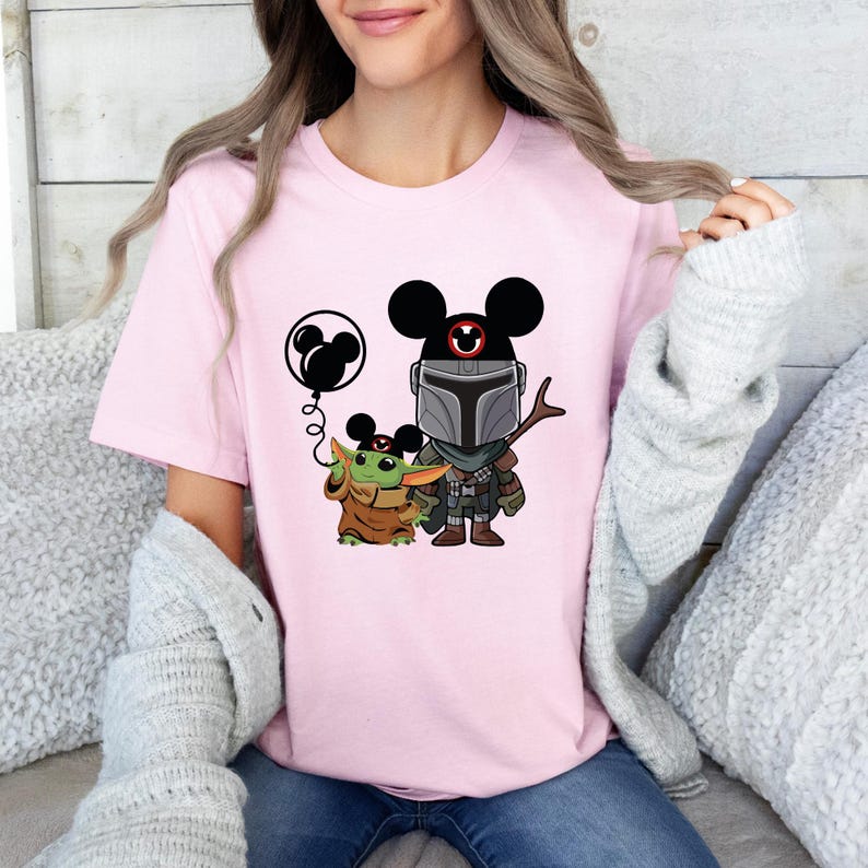 May include: Pink t-shirt with a graphic of a Mandalorian character wearing Mickey Mouse ears and holding a balloon. A small green character with large ears is standing next to him.