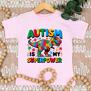 Puede incluir: Camiseta rosa de manga corta con un dinosaurio colorido hecho de piezas de rompecabezas. El texto "El autismo es mi superpoder" est&aacute; impreso en la camiseta.