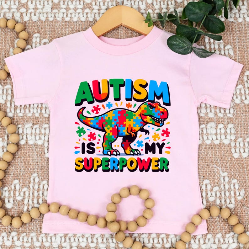 Puede incluir: Camiseta rosa de manga corta con un dinosaurio colorido hecho de piezas de rompecabezas. El texto "El autismo es mi superpoder" est&aacute; impreso en la camiseta.