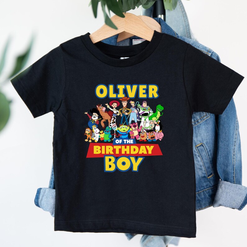May include: Black short-sleeved t-shirt featuring a colorful graphic of animated characters. The text "OLIVER" is above the character group, with "OF THE BIRTHDAY BOY" below. The shirt is hanging on a wooden hanger.