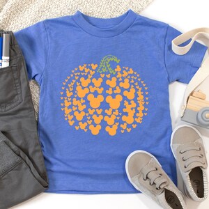 Puede incluir: Una camiseta azul con un dise&ntilde;o de calabaza hecho de siluetas naranjas de Mickey Mouse y un tallo verde. Un pantal&oacute;n y zapatos grises est&aacute;n al lado de la camiseta. Una c&aacute;mara de juguete de madera y una correa blanca tambi&eacute;n est&aacute;n en la imagen.