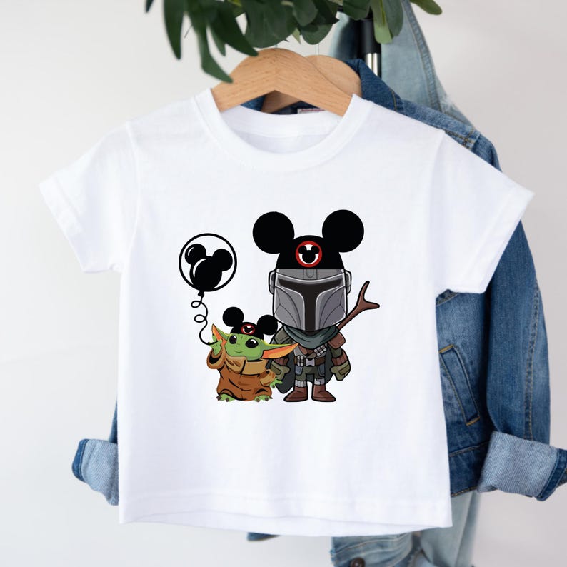 May include: White t-shirt with a cartoon image of a Mandalorian character wearing Mickey Mouse ears and holding a balloon. A small green character with large ears is standing next to him.