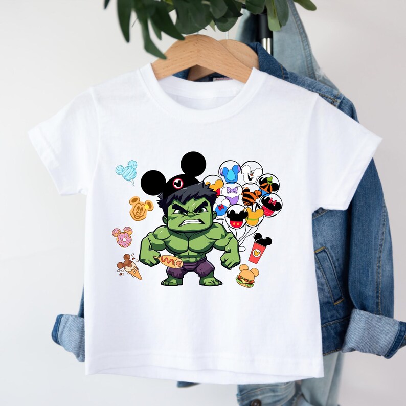 Puede incluir: Camiseta blanca con un personaje verde de Hulk que lleva un sombrero de Mickey Mouse y sostiene un perrito caliente. Hulk est&aacute; rodeado de orejas de Mickey Mouse de colores y globos.