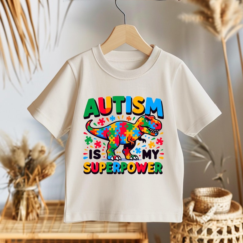 Puede incluir: Una camiseta blanca con un dinosaurio colorido hecho de piezas de rompecabezas. El texto "El autismo es mi superpoder" est&aacute; impreso en la camiseta.