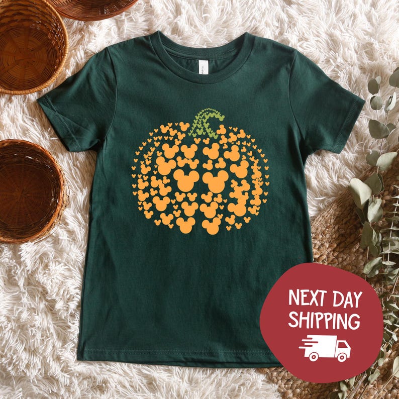 Puede incluir: Camiseta verde oscuro con un dise&ntilde;o de calabaza hecho de siluetas naranjas de Mickey Mouse. La calabaza tiene un tallo verde. La camiseta est&aacute; sobre una superficie blanca y esponjosa. Un c&iacute;rculo rojo con las palabras "NEXT DAY SHIPPING" est&aacute; en la esquina inferior derecha.