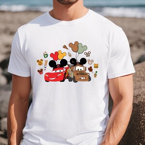 May include: White t-shirt with a colourful graphic of Lightning McQueen and Mater from the Cars film, with Mickey Mouse ears and balloons. The design includes food icons. The shirt is a casual wear item.