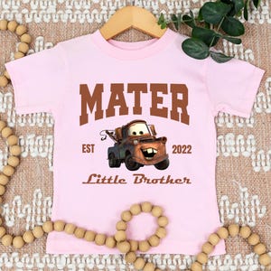 May include: A light pink toddler t-shirt featuring a brown graphic of Mater from the movie Cars. The shirt has the text "MATER", "EST 2022", and "Little Brother" in brown. The shirt is hanging on a wooden hanger.