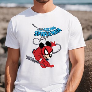 May include: A white short-sleeved t-shirt featuring a graphic print of a character with mouse ears wearing a red and blue spider-themed suit. The text above the character reads "the AMAZING SPIDER-MAN" in blue letters, with black web lines surrounding the figure.