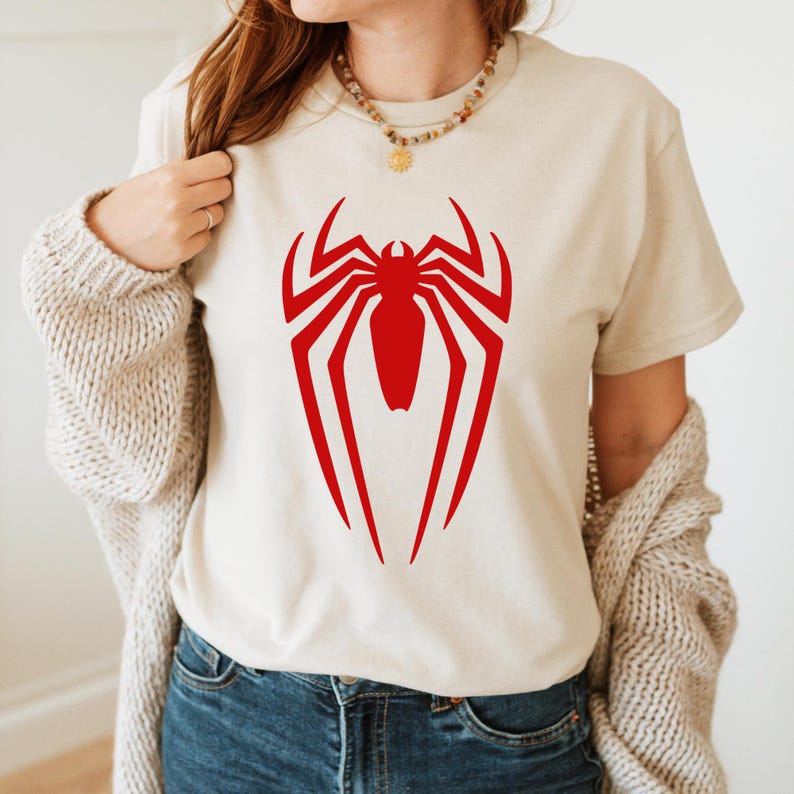May include: A beige t-shirt with a red spider-man logo printed on the front.