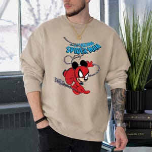 May include: A beige crewneck sweatshirt featuring a graphic print of a character with Mickey Mouse ears wearing a red and blue Spider-Man suit. The text "THE AMAZING SPIDER-MAN" appears in blue above the character, who is swinging on a web.