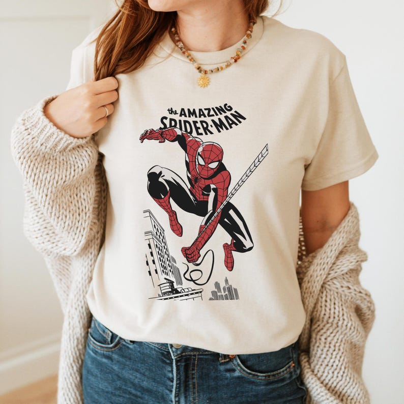 May include: A beige t-shirt with a graphic print of Spider-Man swinging through the city. The text "The Amazing Spider-Man" is printed above the image.