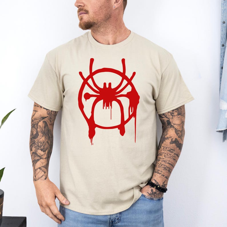 Puede incluir: Una camiseta beige con un s&iacute;mbolo de Spiderman rojo. El s&iacute;mbolo es una ara&ntilde;a estilizada con un c&iacute;rculo alrededor, y el c&iacute;rculo tiene gotas de pintura roja que salen de &eacute;l.