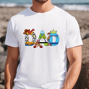 K&ouml;nnte beinhalten: Wei&szlig;es T-Shirt mit einer Grafik des Wortes "DAD", das aus Figuren aus dem Toy Story-Film-Franchise besteht. Die Figuren sind Woody, Buzz Lightyear und der Au&szlig;erirdische.