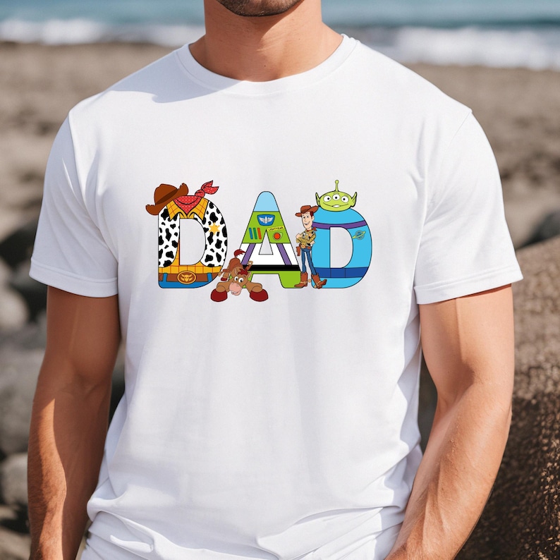 K&ouml;nnte beinhalten: Wei&szlig;es T-Shirt mit einer Grafik des Wortes "DAD", das aus Figuren aus dem Toy Story-Film-Franchise besteht. Die Figuren sind Woody, Buzz Lightyear und der Au&szlig;erirdische.