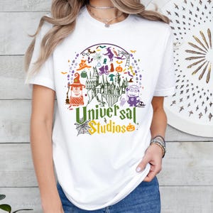 May include: White t-shirt featuring a colorful graphic design with the text "Universal Studios". The design includes Halloween-themed elements such as a witch, a haunted castle, a minion, and a pumpkin.