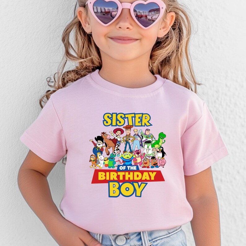 May include: A light pink t-shirt featuring a colorful graphic of characters from the Toy Story movie. The text "SISTER OF THE BIRTHDAY BOY" is displayed in yellow and red. The shirt is short-sleeved.