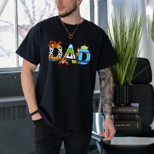 K&ouml;nnte beinhalten: Schwarzes T-Shirt mit einem Aufdruck von Figuren aus dem Film Toy Story, die "DAD" buchstabieren.