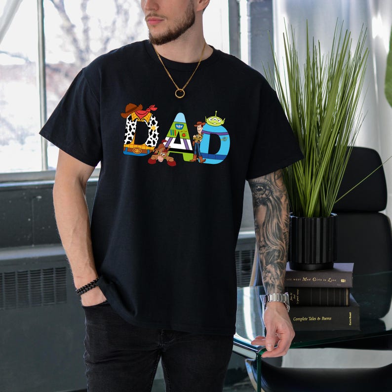 K&ouml;nnte beinhalten: Schwarzes T-Shirt mit einem Aufdruck von Figuren aus dem Film Toy Story, die "DAD" buchstabieren.