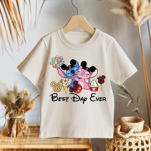 May include: Cream-colored t-shirt featuring a graphic of two cartoon characters hugging, with the text "Best Day Ever" in black. The graphic includes colorful balloons, ice cream, and other treats. The shirt is hanging on a wooden hanger.