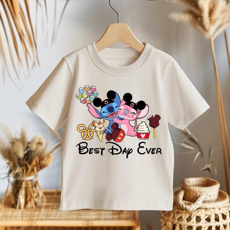 May include: Cream-colored t-shirt featuring a graphic of two cartoon characters hugging, with the text "Best Day Ever" in black. The graphic includes colorful balloons, ice cream, and other treats. The shirt is hanging on a wooden hanger.