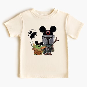 May include: A cream-colored t-shirt with a black and green cartoon image of a Mandalorian and a Grogu character wearing Mickey Mouse ears. The Mandalorian is holding a stick and Grogu is holding a black balloon with a Mickey Mouse head.