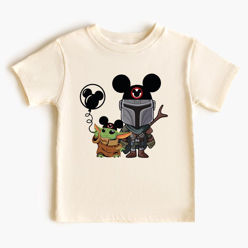 May include: A cream-colored t-shirt with a black and green cartoon image of a Mandalorian and a Grogu character wearing Mickey Mouse ears. The Mandalorian is holding a stick and Grogu is holding a black balloon with a Mickey Mouse head.
