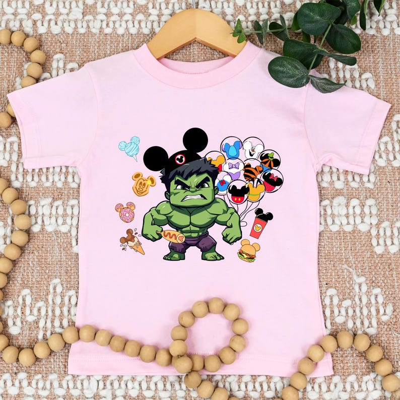 Puede incluir: Camiseta rosa de manga corta con un personaje verde de Hulk que lleva orejas de Mickey Mouse. Hulk sostiene un perrito caliente y est&aacute; rodeado de globos coloridos y otras im&aacute;genes con tem&aacute;tica de Disney.