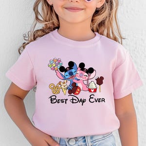 May include: Pink t-shirt featuring a graphic of two cartoon characters hugging, with ice cream and the text "Best Day Ever." The characters are blue and pink, with black ears. The shirt is for children.