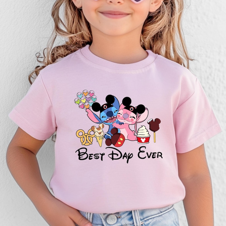 May include: Pink t-shirt featuring a graphic of two cartoon characters hugging, with ice cream and the text "Best Day Ever." The characters are blue and pink, with black ears. The shirt is for children.