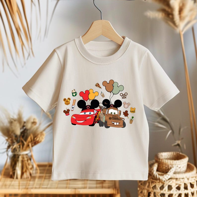 May include: Cream-coloured short-sleeved t-shirt featuring a colourful graphic of two cartoon cars with Mickey Mouse ears and balloons. The design includes various food and character icons. The shirt is hanging on a wooden hanger.
