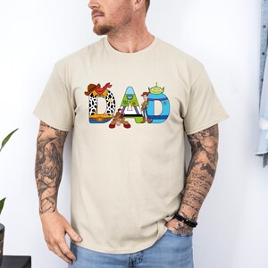 K&ouml;nnte beinhalten: Ein beiges T-Shirt mit einer Grafik des Wortes "DAD", das aus Figuren aus dem Toy Story-Film-Franchise besteht. Die Figuren sind Woody, Buzz Lightyear und der Au&szlig;erirdische.