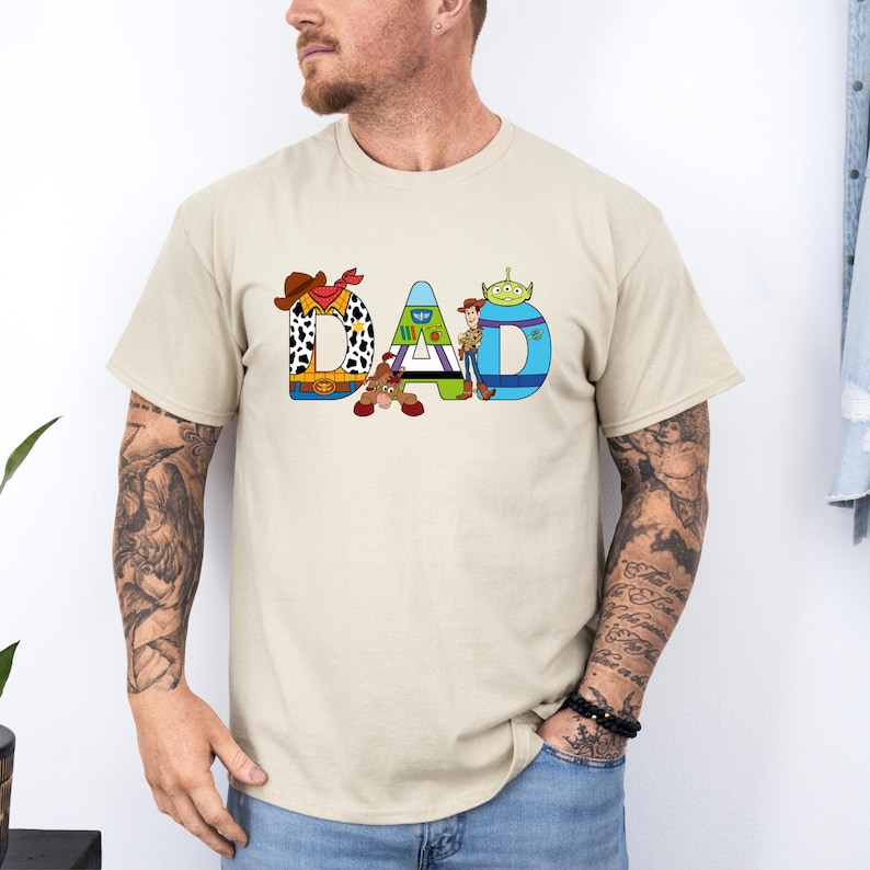 K&ouml;nnte beinhalten: Ein beiges T-Shirt mit einer Grafik des Wortes "DAD", das aus Figuren aus dem Toy Story-Film-Franchise besteht. Die Figuren sind Woody, Buzz Lightyear und der Au&szlig;erirdische.