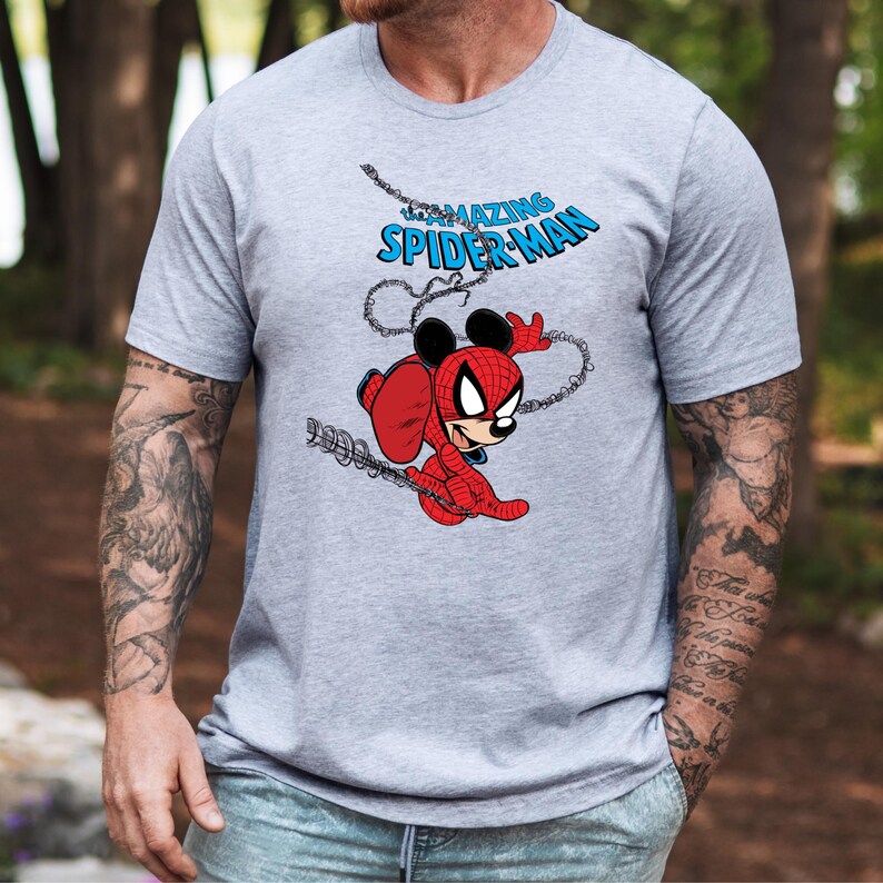 May include: A heather gray short-sleeve t-shirt featuring a graphic print of a character wearing a red and black spider-themed suit with mouse ears. Above the character, the text reads THE AMAZING SPIDER-MAN in blue letters with a web design.