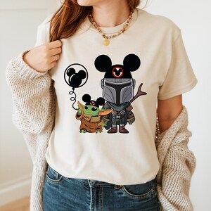 May include: A white t-shirt with a black and green cartoon image of a Mandalorian and a Grogu character wearing Mickey Mouse ears. The Mandalorian is holding a stick and Grogu is holding a balloon with Mickey Mouse ears.