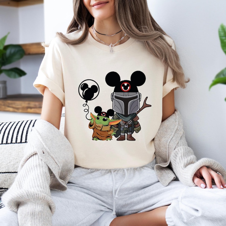 May include: A white t-shirt with a graphic of a Mandalorian character wearing Mickey Mouse ears and holding a balloon. The Mandalorian is standing next to a small green character wearing Mickey Mouse ears. The text "This is the way" is printed on the shirt.