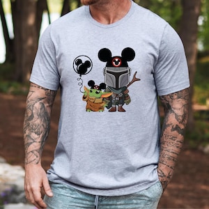 May include: A gray t-shirt with a cartoon image of a Mandalorian and Grogu wearing Mickey Mouse ears. Grogu is holding a balloon shaped like Mickey Mouse's head.