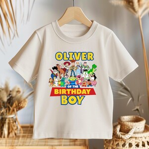 May include: Off-white short-sleeved t-shirt featuring a colorful graphic of animated characters. The text "OLIVER" is above the characters, and "OF THE BIRTHDAY BOY" is below. The shirt is hanging on a wooden hanger.
