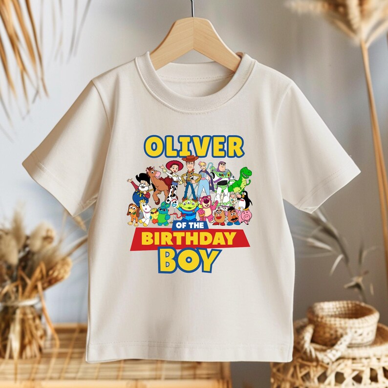 May include: Off-white short-sleeved t-shirt featuring a colorful graphic of animated characters. The text "OLIVER" is above the characters, and "OF THE BIRTHDAY BOY" is below. The shirt is hanging on a wooden hanger.