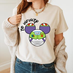 May include: A beige t-shirt with a graphic of Buzz Lightyear's helmet in the shape of Mickey Mouse ears. The text "To Infinity" is written above the graphic.