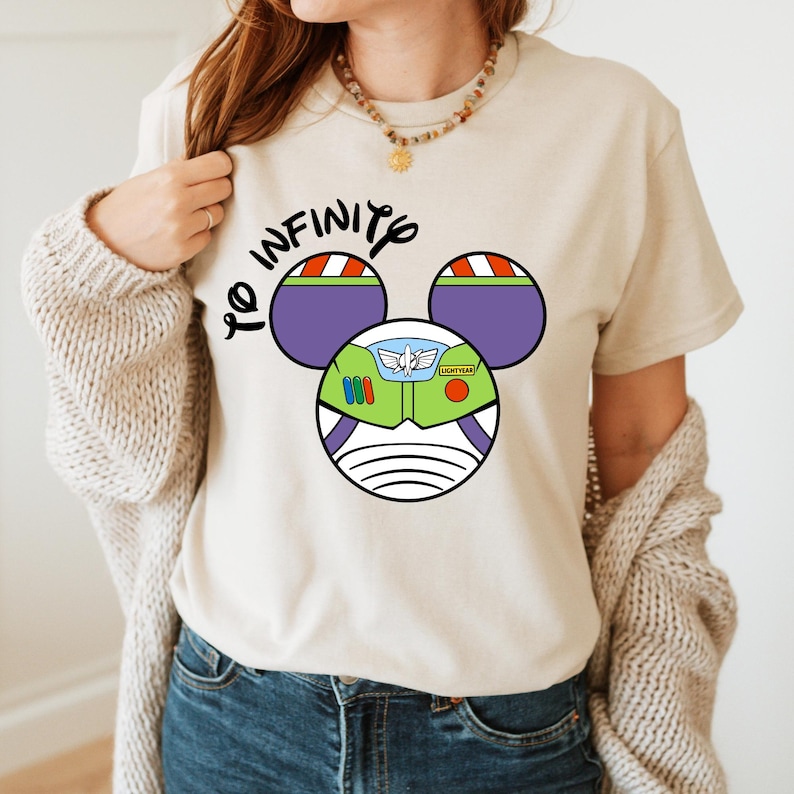 May include: A beige t-shirt with a graphic of Buzz Lightyear's helmet in the shape of Mickey Mouse ears. The text "To Infinity" is written above the graphic.