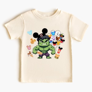 Puede incluir: Una camiseta de color crema claro con una imagen de dibujos animados de Hulk con un sombrero de Mickey Mouse y sosteniendo un perrito caliente. Hulk est&aacute; rodeado de im&aacute;genes de dibujos animados de orejas de Mickey Mouse, globos y comida.