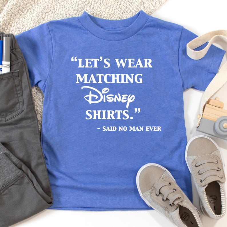 Peut inclure: Un t-shirt bleu avec le texte "LET'S WEAR MATCHING Disney SHIRTS." et "- SAID NO MAN EVER" en blanc. Le t-shirt est associ&eacute; &agrave; un pantalon gris et des baskets. Un petit appareil photo jouet et une sangle cr&egrave;me sont &eacute;galement visibles.