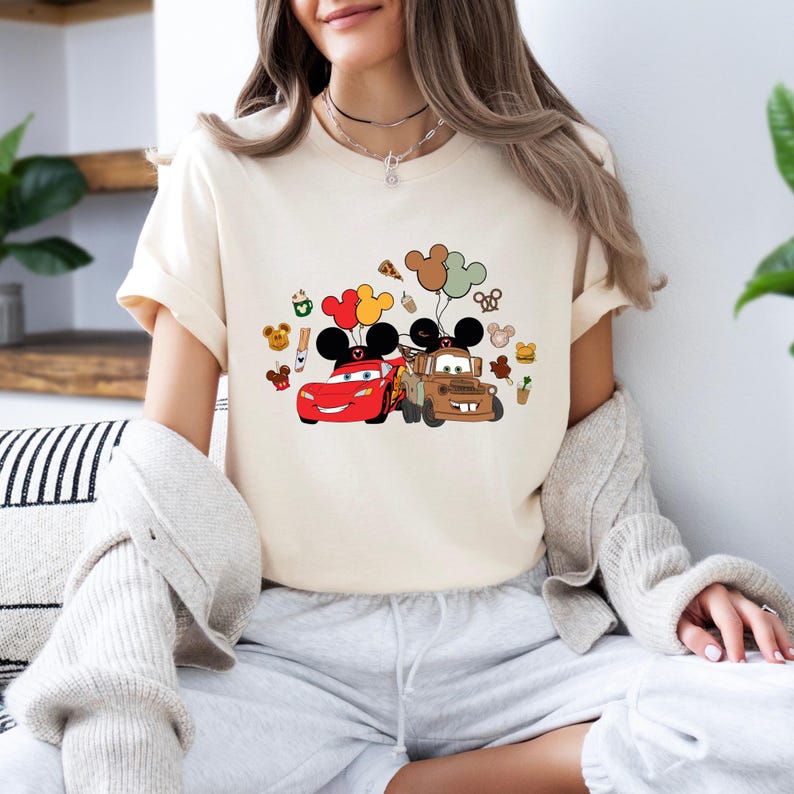 May include: A cream-coloured t-shirt with a graphic of the Cars characters, Lightning McQueen and Mater, alongside Mickey Mouse balloons and other Disney-themed icons. The design incorporates red, yellow, brown, and black colours.
