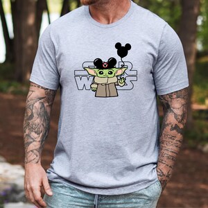 May include: A grey t-shirt featuring a green character wearing a Mickey Mouse hat and holding a black balloon. The text "STAR WARS" is printed on the shirt.