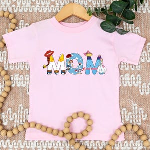 K&ouml;nnte beinhalten: Rosa T-Shirt mit kurzen &Auml;rmeln, auf dem das Wort "MOM" in bunten Buchstaben geschrieben ist, die Figuren aus dem Toy Story-Franchise darstellen.