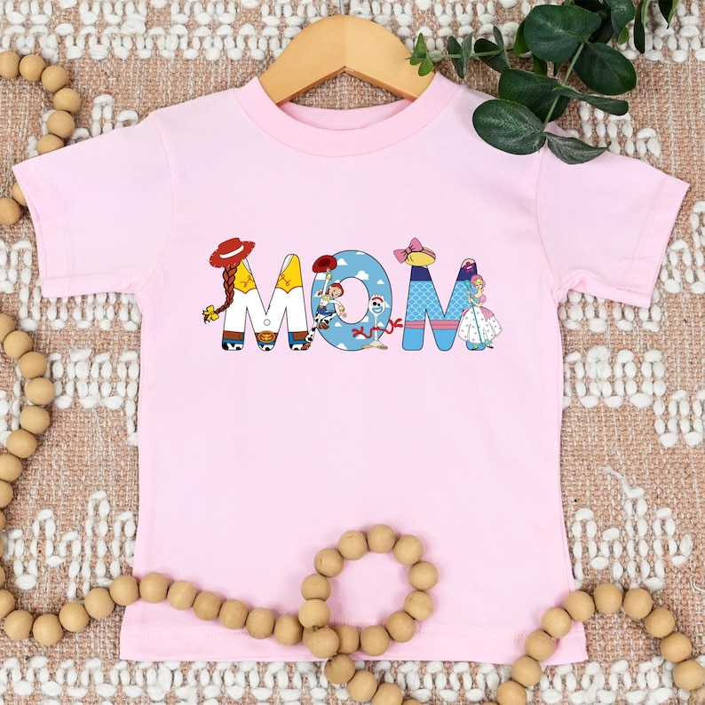 K&ouml;nnte beinhalten: Rosa T-Shirt mit kurzen &Auml;rmeln, auf dem das Wort "MOM" in bunten Buchstaben geschrieben ist, die Figuren aus dem Toy Story-Franchise darstellen.