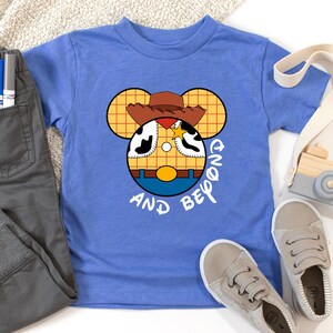 May include: A blue t-shirt with a cartoon image of Woody from Toy Story in a Mickey Mouse silhouette. The text "And Beyond" is printed below the image.