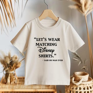 Peut inclure: Un t-shirt cr&egrave;me avec le texte "LET'S WEAR MATCHING Disney SHIRTS." et "- SAID NO MAN EVER" en noir. Le t-shirt est suspendu &agrave; un cintre en bois.