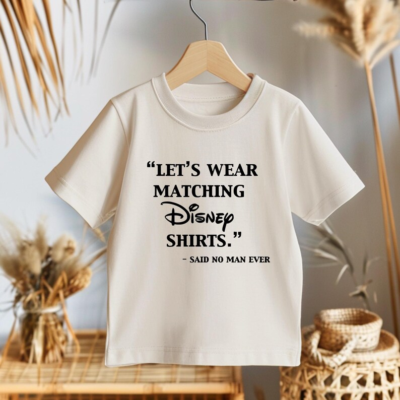 Peut inclure: Un t-shirt cr&egrave;me avec le texte "LET'S WEAR MATCHING Disney SHIRTS." et "- SAID NO MAN EVER" en noir. Le t-shirt est suspendu &agrave; un cintre en bois.