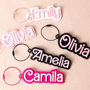 Op de afbeelding: Vijf gepersonaliseerde naamsleutelhangers met gelaagde acryldesigns. De sleutelhangers tonen de namen Emily, Olivia, Amelia en Camila in diverse kleurcombinaties, waaronder roze, wit en zwart, elk bevestigd aan een ronde metalen ring.